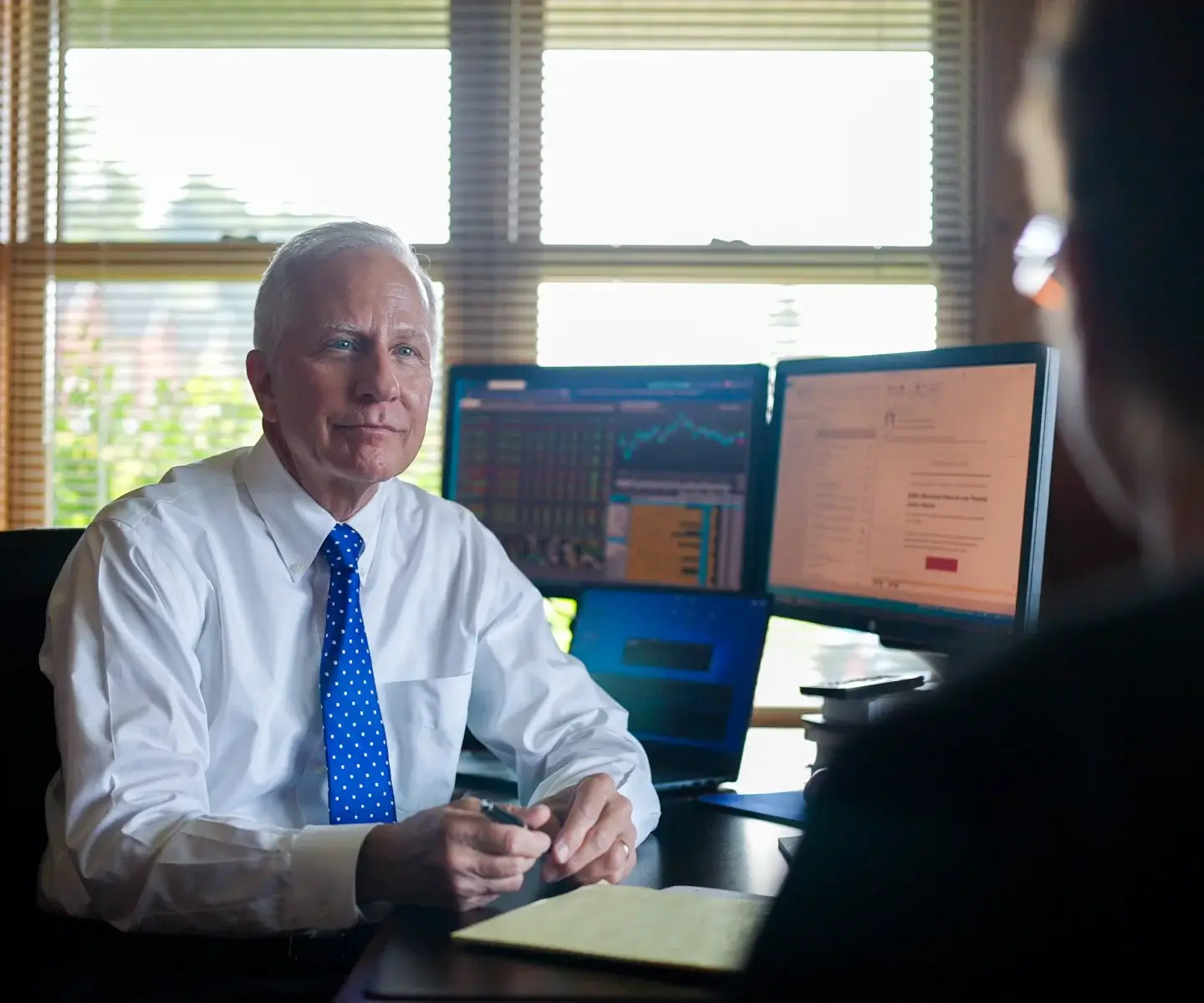 An older man in a white shirt and blue tie sits at a desk with multiple computer monitors, facing another person.